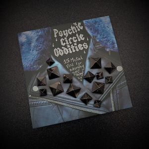May include: A set of 15 black metal pyramid-shaped pins, displayed on a blue denim jacket with faux fur collar. The text "Psychic Circle Oddities" and "15 Metal Pins for Adorning your things xx" is written on the jacket.