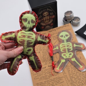 May include: A green and brown felt skeleton doll with red stitching. The doll is being held by a hand. The doll is part of a craft kit that includes sewing instructions and herbs.