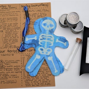May include: A blue skeleton witch poppet kit with instructions, herbs, and a small glass vial. The kit includes a blue skeleton witch poppet, a spool of blue thread, four small jars of herbs, a small glass vial, and a black box with a white paper insert. The instructions include information about the herbs and how to use the poppet.