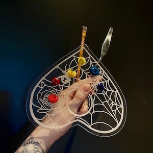 May include: A clear plastic paint palette shaped like a heart with a spiderweb design. The palette has five paint wells filled with red, yellow, blue, and green paint. Two paintbrushes are resting on the palette.