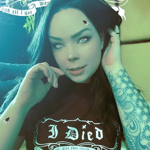 May include: A black t-shirt with the phrase "I Died and all I got was this lousy t-shirt" in a gothic font. The image shows a person with dark hair and a tattoo on their arm. The person is wearing the t-shirt.