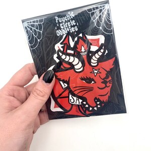 May include: A set of three stickers featuring a red and black illustration of a cat with horns and a pentagram. The stickers are packaged in a clear plastic bag with a black and white spiderweb design. The text "Psychic Circle Oddities" is printed on the bag.