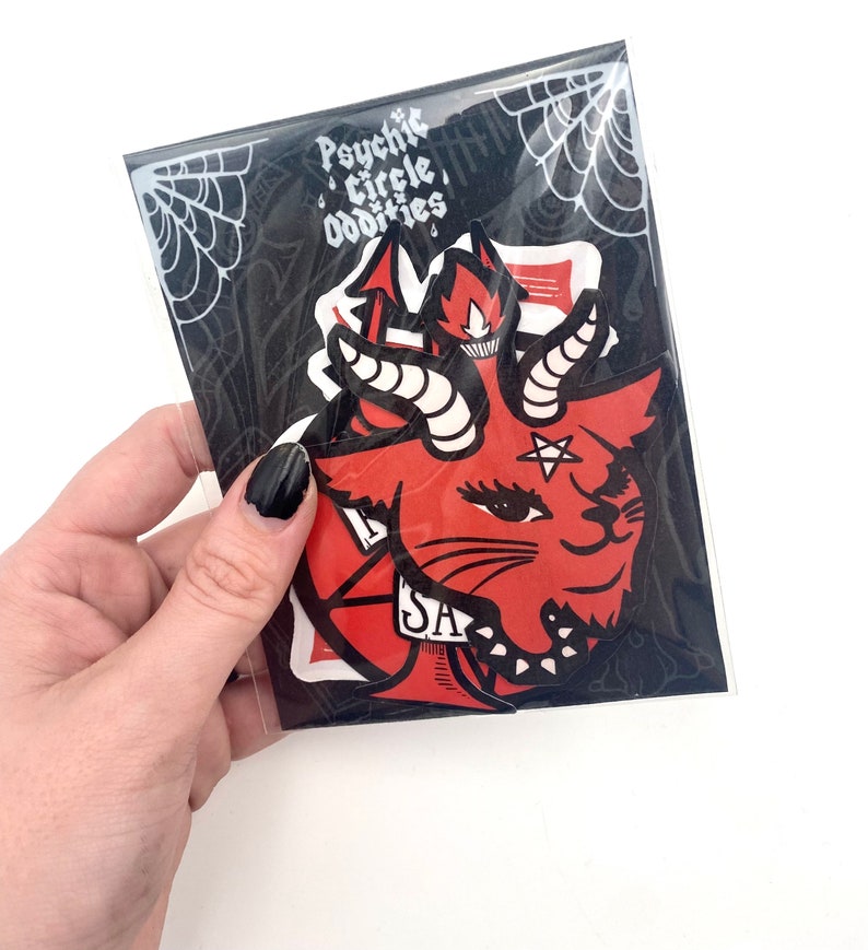 May include: A set of three stickers featuring a red and black illustration of a cat with horns and a pentagram. The stickers are packaged in a clear plastic bag with a black and white spiderweb design. The text "Psychic Circle Oddities" is printed on the bag.