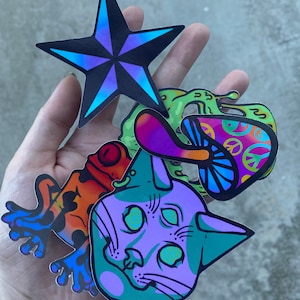 May include: A set of five colorful stickers featuring a blue star, a green and purple mushroom, a green and purple cat with spiral eyes, a green and orange frog, and a green and purple peace sign.