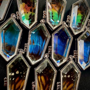 May include: A silver necklace featuring multiple crystal-like pendants, each containing a preserved butterfly wing. The wings are in various colours, including blue, green, and brown.