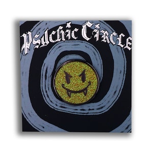 May include: A green glitter enamel pin with a black outline of a smiley face with fangs. The pin is on a black and blue background with the text "Psychic Circle" in black.