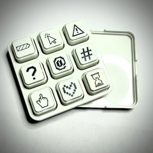 May include: A white, square-shaped keypad with nine buttons, each displaying a black pixelated icon. Icons include a battery, mouse cursor, exclamation point, question mark, at symbol, hashtag, middle finger, heart, and hourglass. The keypad is next to a white, rectangular case.
