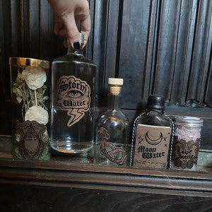 May include: Four glass bottles with labels and a small jar filled with pink crystals. The bottles are labeled "Storm Water", "Made So", "Moon Water", and "Love Bottle".