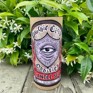 Pode incluir: Um conjunto de quatro velas de soja de 3 oz em uma caixa de papel&atilde;o marrom com uma ilustra&ccedil;&atilde;o em preto e branco de um olho com uma borda vermelha e branca. O texto na caixa diz "Psychic Circle Oddities Candle Set".