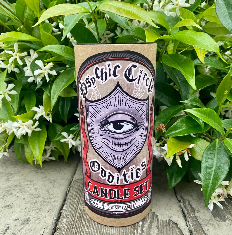 Pode incluir: Um conjunto de quatro velas de soja de 3 oz em uma caixa de papel&atilde;o marrom com uma ilustra&ccedil;&atilde;o em preto e branco de um olho com uma borda vermelha e branca. O texto na caixa diz "Psychic Circle Oddities Candle Set".