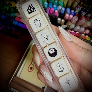May include: A collection of small, square stamps featuring black and white illustrations: flames, a tooth, sparkles, an eight ball, an anchor, and a dagger. The stamps are housed in a clear plastic case, with a metal tin visible.