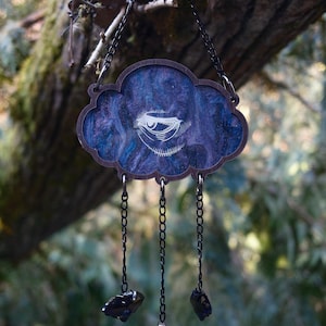 May include: A dark blue and purple resin cloud-shaped ornament with a white eye design. The ornament hangs from a black chain with three dangling charms: two black stones and a iridescent shell.