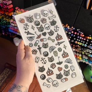 May include: A rectangular clear plastic sheet containing a collection of small, individual stickers featuring various illustrations including black cats, skulls, bones, anchors, dice, eight balls, flames, spiderwebs, hearts, and daggers.