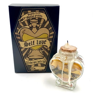 May include: A black box with gold lettering that reads "Psychic Circle Oddities presents Self Love Witches Spell Bottle Kit". The box contains a heart-shaped glass bottle with a cork stopper and a white candle. The bottle is filled with various herbs and crystals.