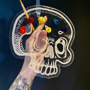Può includere: Una tavolozza di vernice in plastica trasparente a forma di teschio con un contorno bianco. La tavolozza ha cinque pozzetti di vernice riempiti di vernice rossa, gialla, blu e verde. Un pennello di legno è appoggiato sulla tavolozza.