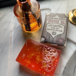 May include: A bar of red soap with gold flakes and white bubbles. The soap is labelled "Psychic Circle Oddities Thieves Oil Based on an Ancient Medieval Medicinal Recipe Luxury Soap Handcrafted with Magic Home is Portland Oregon"