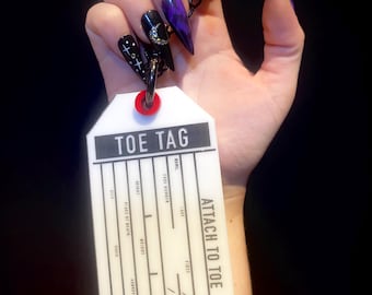 May include: A white toe tag with the text "TOE TAG" and "ATTACH TO TOE" in black. The tag is held by a hand with long, pointed, black and purple nails. The nails are decorated with stars, moons, and glitter.
