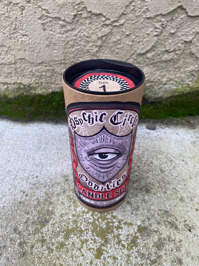 Pode incluir: Um tubo de cart&atilde;o castanho com uma tampa preta. O tubo tem um r&oacute;tulo com uma ilustra&ccedil;&atilde;o em preto e branco de um olho dentro de um escudo. O r&oacute;tulo tamb&eacute;m diz "Psychic Circle Oddities Candle Set" e "4 - 4 oz Soy Candles".