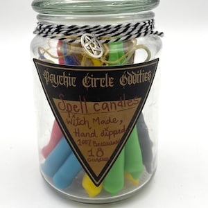 Pode incluir: Um frasco de vidro transparente cheio de velas de feitiço coloridas. O frasco está rotulado como "Psychic Circle Oddities" e "Spell candles". As velas são mergulhadas à mão, feitas com 100% de cera de abelha, e há 18 velas no frasco. O frasco é amarrado com barbante preto e branco e um pingente de prata.