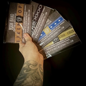 May include: A collection of elastic book bands in clear plastic packaging, each featuring unique designs like wood grain, chains, blue stars, and yellow caution tape. The packaging includes the text BOOK BAND and WWW.PSYCHICCIRCLEODDITIES.COM.