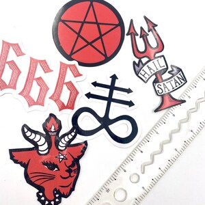 May include: A set of six red and black stickers with various satanic symbols, including a pentagram, an upside-down cross, a trident, and the number 666. The stickers are arranged on a white surface with a ruler.