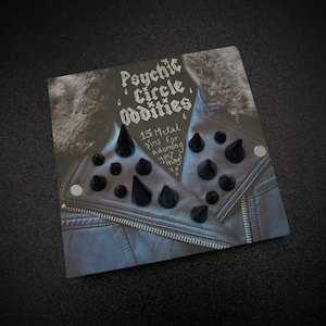 May include: A set of 15 black metal pins with pointed tips, displayed on a black leather jacket. The pins are for adorning your things. The text "Psychic Circle Oddities" and "15 Metal Pins for Adorning Your Things" is written on the card.