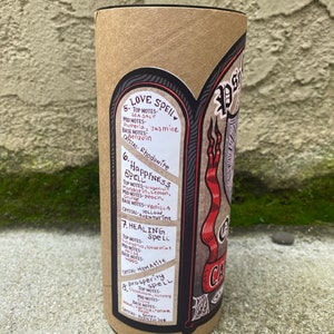 Pu&ograve; includere: Un tubo di cartone marrone con un'etichetta nera e rossa. L'etichetta presenta un'illustrazione in bianco e nero di una ragnatela e un nastro rosso con il testo "Candle Spell". L'etichetta include anche un elenco di diversi incantesimi con le loro note di cristallo e profumo corrispondenti. Gli incantesimi includono Love Spell, Happiness Spell, Healing Spell e Prosperity Spell.