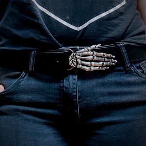 May include: Black leather belt with a silver-colored skeleton hand buckle. The belt is worn with dark wash denim jeans and a black t-shirt. The buckle is the focal point, adding a unique, gothic touch to the outfit.