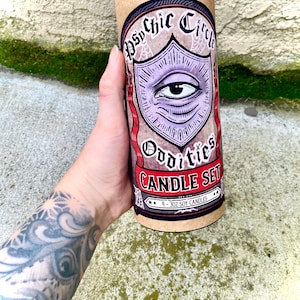 Pu&ograve; includere: Un tubo di cartone marrone con un coperchio nero. Il tubo presenta un design grafico di un occhio viola con uno striscione rosso che dice "Psychic Circle Oddities Candle Set". Il testo "4 - 3 oz Soy Candles" &egrave; stampato sul tubo.