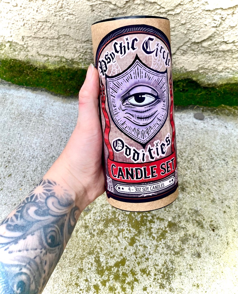 Pu&ograve; includere: Un tubo di cartone marrone con un coperchio nero. Il tubo presenta un design grafico di un occhio viola con uno striscione rosso che dice "Psychic Circle Oddities Candle Set". Il testo "4 - 3 oz Soy Candles" &egrave; stampato sul tubo.