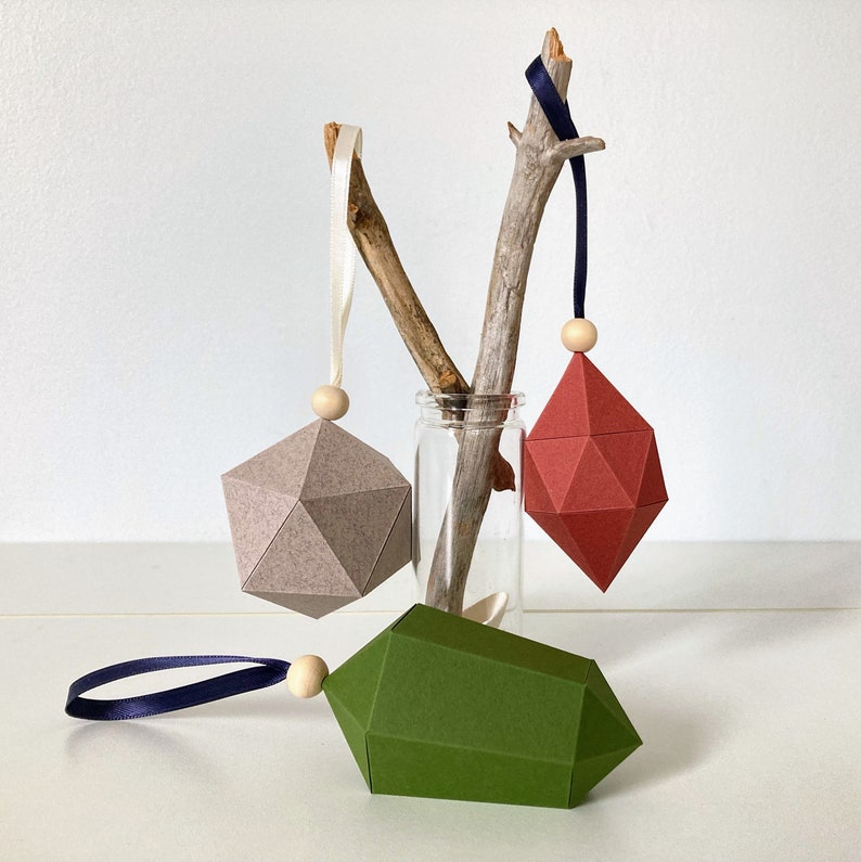 One speckled gray and one clay colored geometric ornament hang from sticks of driftwood. A moss green geometric ornament lies in front.