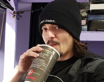 May include: A person wearing a black beanie with the text "Staff Cowboy" on it, a black jacket, a black t-shirt with the text "Geek Squad" on it, and blue jeans. The person is holding a black and white cup with a red logo and text.