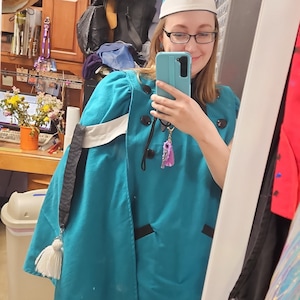 Op de afbeelding: Een persoon draagt een turquoise kostuum met een witte kraag en zwarte bies. Het kostuum heeft een witte band en een zwarte kwast. De persoon draagt een hoge, puntige hoed met een gouden rand en een witte rand. De hoed heeft een gouden kwast bovenop. De persoon houdt een mobiele telefoon in zijn rechterhand en kijkt naar de camera.