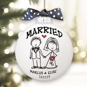 May include: A white spherical Christmas ornament featuring a black line drawing of a couple, the word MARRIED, a small red heart, and the names MARCUS & ELISE with the date 10.22.23. It is topped with a dark blue ribbon with white polka dots.