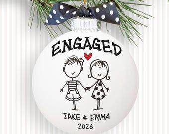 May include: A white spherical Christmas ornament featuring the black text ENGAGED, a red heart, and a line drawing of two people holding hands. Below the figures, the names JAKE & EMMA and the year 2026 are printed. A black and white polka dot bow sits on top.