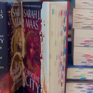 May include: A stack of books, with the spine of a book by Sarah J. Maas in focus. The book is titled "Crescent City" and has a red and gold cover. The pages are marked with colorful sticky notes.
