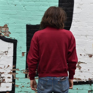 May include: A person wearing a red sweatshirt and blue jeans standing in front of a white brick wall with green and black graffiti.