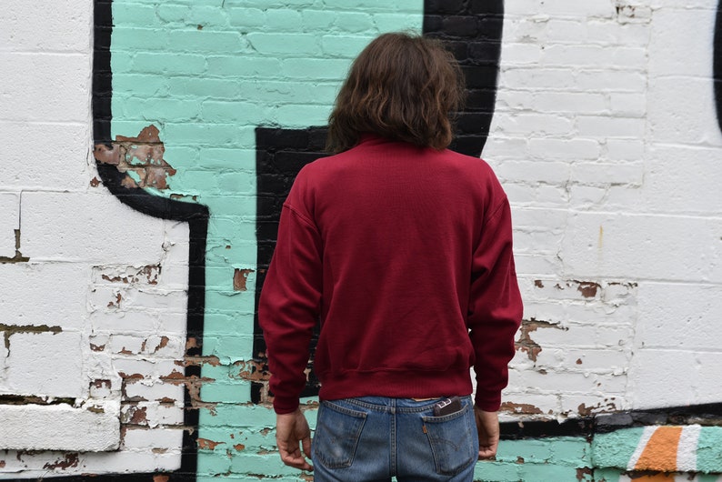 May include: A person wearing a red sweatshirt and blue jeans standing in front of a white brick wall with green and black graffiti.