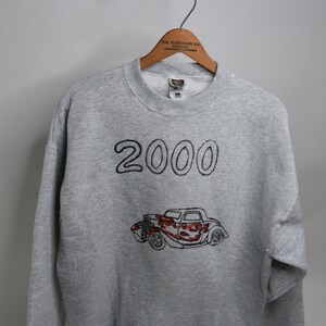 Op de afbeelding: Grijs sweatshirt met een zwart geborduurd ontwerp van een hotrod-auto en het jaar 2000.