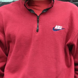 May include: A red Nike sweatshirt with a quarter-zip closure and a blue and white Nike logo on the chest. The logo says "NIKE" in white letters.