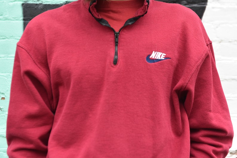 May include: A red Nike sweatshirt with a quarter-zip closure and a blue and white Nike logo on the chest. The logo says "NIKE" in white letters.