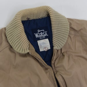 May include: A tan jacket with a brown knit collar and a blue lining. The jacket has a zipper closure and a label that reads "Woolrich Est. 1830 Made in USA".
