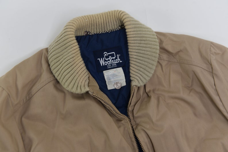 May include: A tan jacket with a brown knit collar and a blue lining. The jacket has a zipper closure and a label that reads "Woolrich Est. 1830 Made in USA".
