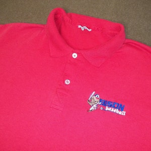 Può includere: Polo rosso a maniche corte con un logo ricamato di un giocatore di baseball e il testo "BISON baseball".