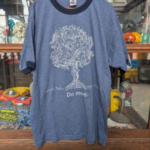 May include: A blue t-shirt with a white graphic of a tree with the text "Do more." printed on the front. The shirt has a black ribbed crew neck.