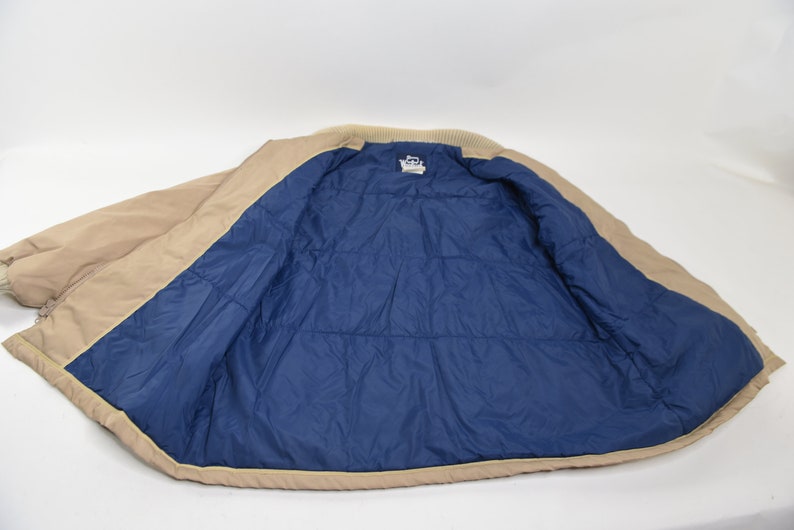 May include: A tan puffer jacket with a blue quilted lining. The jacket is unzipped and laid flat.