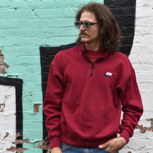 May include: A red Nike sweatshirt with a half-zip closure and a small Nike logo on the left chest. The sweatshirt is worn by a person with long hair and glasses.