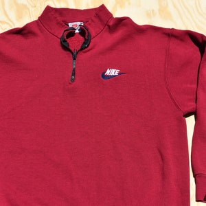 May include: A red Nike quarter-zip sweatshirt with a navy blue swoosh logo and the word "NIKE" embroidered on the front.
