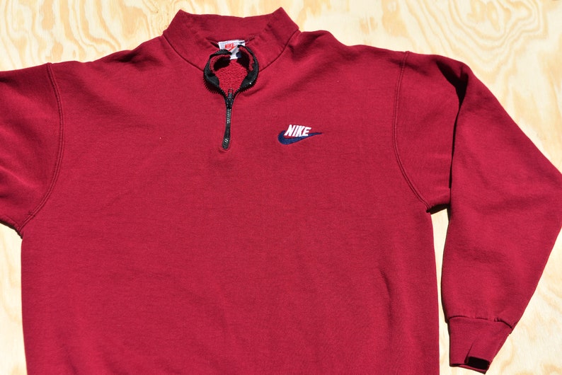 May include: A red Nike quarter-zip sweatshirt with a navy blue swoosh logo and the word "NIKE" embroidered on the front.
