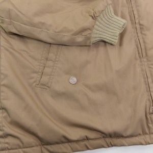 May include: A tan jacket with a zipper closure and a ribbed knit cuff. The jacket has a pocket on the left side and a zipper pull with a blue tab.